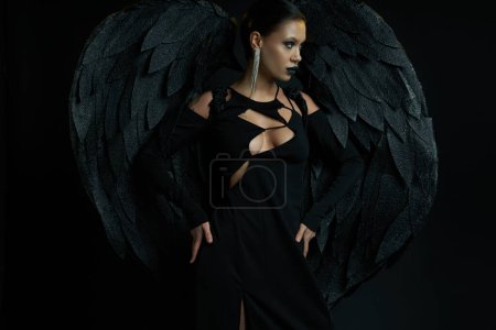 mujer en maquillaje oscuro y traje de fantasía de criatura alada demoníaca mirando hacia otro lado en negro