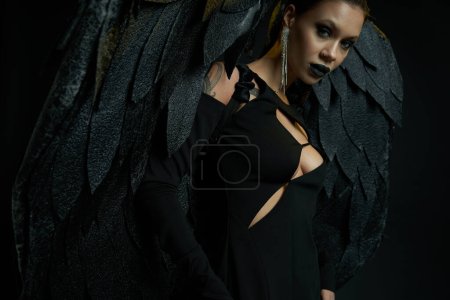 verführerische tätowierte Frau im Halloween-Kostüm eines dunklen Dämons mit Flügeln, die auf schwarz in die Kamera schaut