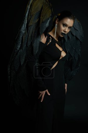 sexy tattooed woman in halloween costume of fallen angel with dark wings looking at camera on black magic mug #676127420
