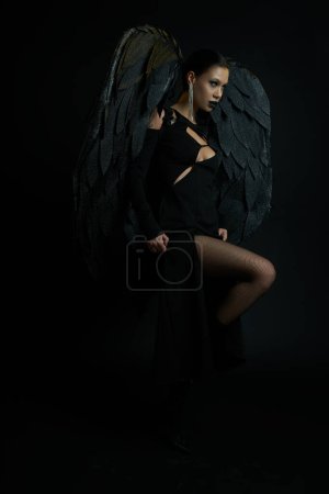 Foto de Seductora mujer en maquillaje oscuro y disfraz con alas demoníacas mirando hacia otro lado en negro, Halloween - Imagen libre de derechos