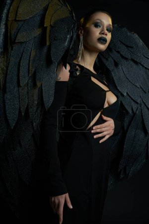 dark beauty, tattooed woman in halloween costume of winged fallen angel looking at camera on black
