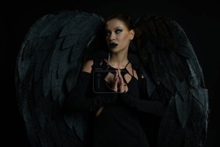 mujer tatuada en traje de ángel oscuro de pie con las manos de oración y mirando hacia otro lado en negro