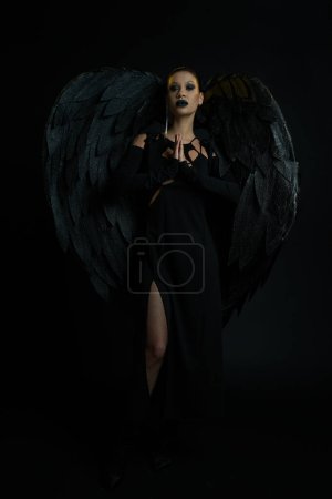 mysterious woman in costume of winged creature standing with praying hands on black, demonic beauty