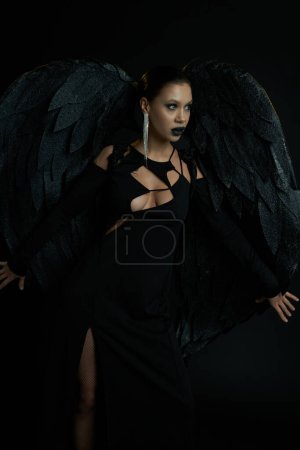 séduisante femme en costume de démon sombre avec des ailes regardant loin sur noir, concept halloween