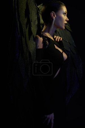 Photo for Demonic charm, side view of sexy woman in halloween costume of dark demon with wings on black - Royalty Free Image