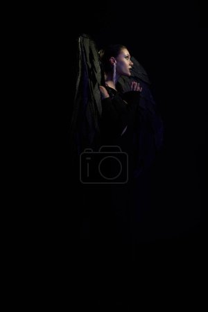 boczny widok czarującej kobiety jako ciemnego demona ze skrzydłami modlącymi się na czarnym tle, koncepcja halloween