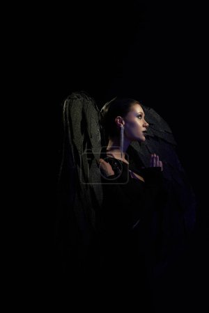 Foto de Vista lateral de la mujer en traje de ángel caído con alas oscuras rezando en la oscuridad, fondo negro - Imagen libre de derechos