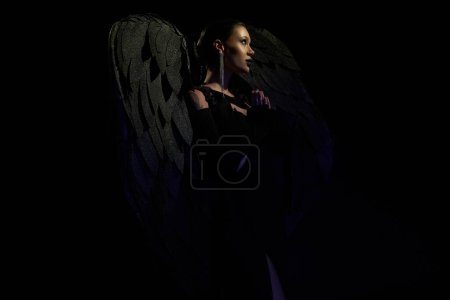 widok z boku tajemniczej kobiety w kostiumie demonicznej skrzydlatej istoty modlącej się na czarnym tle