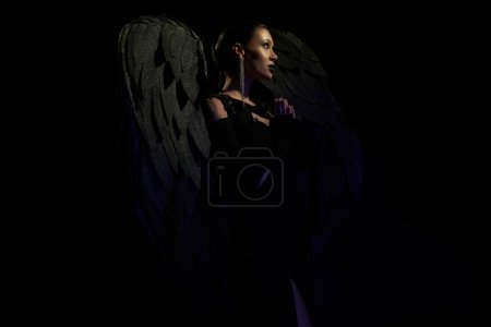 Foto de Vista lateral de mujer misteriosa disfrazada de criatura alada demoníaca rezando sobre fondo negro - Imagen libre de derechos