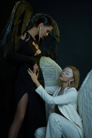 women in Halloween costumes, white angel sitting on haunches near dark demon on black backdrop