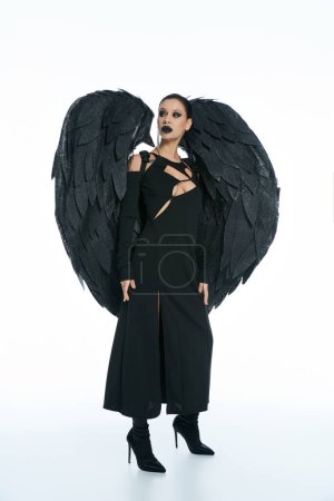 longitud completa de la misteriosa mujer en traje de demonio alado negro mirando hacia otro lado en el fondo blanco