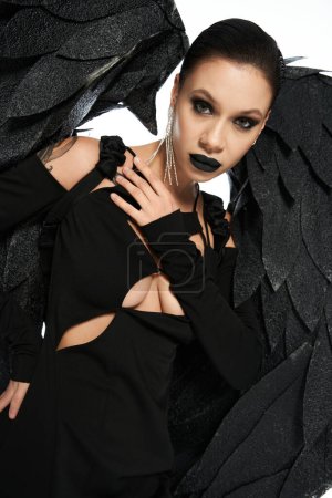 Foto de Mujer encantadora en maquillaje oscuro y traje de criatura alada negro mirando a la cámara en blanco - Imagen libre de derechos