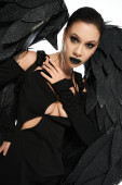 enchanting woman in dark makeup and costume of black winged creature looking at camera on white Sweatshirt #676128026
