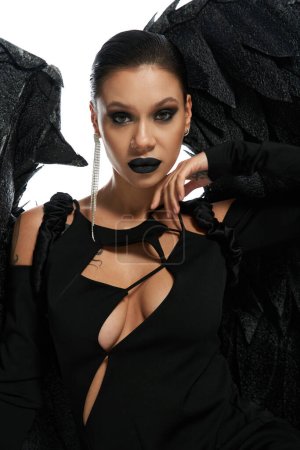 retrato de mujer sexy en maquillaje oscuro y traje de demonio alado negro mirando a la cámara en blanco