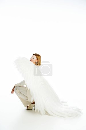 full length of woman in costume of light angel with heavenly wings sitting on haunches on white