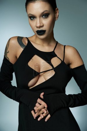 verführerisch tätowierte Frau mit dunklem Make-up trägt schwarzes sexy Kleid und schaut in die Kamera auf grau