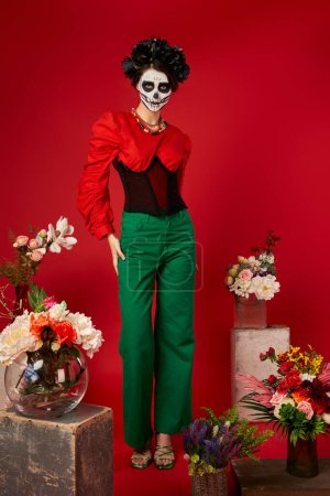 Photo for Woman in sugar skull makeup near traditional dia de los muertos ofrenda with bright flowers on red - Royalty Free Image