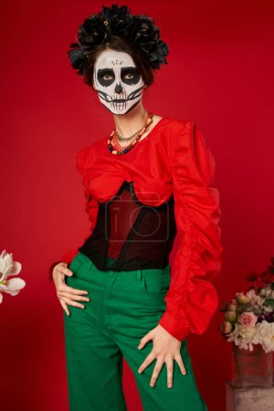 woman in sugar skull makeup and black wreath near flowers on red, dia de los muertos tradition