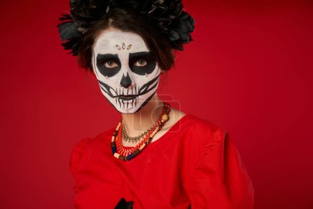 portrait of woman in sugar skull makeup and black wreath looking at camera on red, Day of Dead