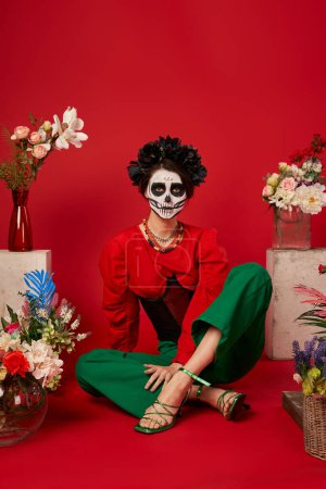 Frau in Catrina Make-up sitzt in der Nähe der traditionellen dia de los muertos ofrenda mit Blumen auf rot
