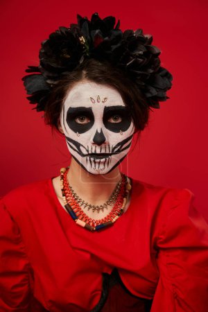 woman in dia de los muertos makeup and black wreath with colorful beads looking at camera on red