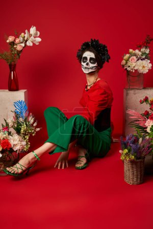 woman in skull makeup and black wreath sitting near dia de los muertos ofrenda with flowers on red