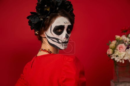 portrait of young woman in spooky catrina makeup and black wreath looking at camera on red