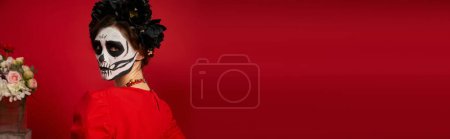 Frau in Totenkopf-Make-up blickt in die Kamera neben bunten Blumen auf rot, dia de los muertos, Banner