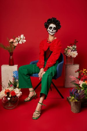 Frau in dia de los muertos Make-up sitzt im Sessel in der Nähe der traditionellen ofrenda mit Blumen auf rot
