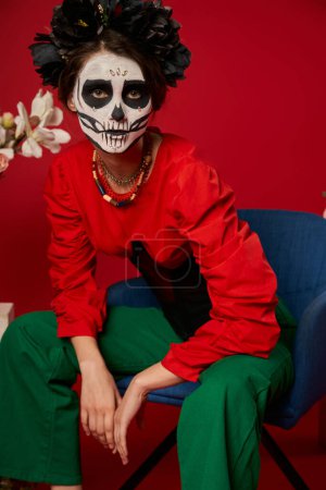 Frau in dia de los muertos Make-up sitzt im Sessel neben Blumen und blickt in die Kamera auf rot