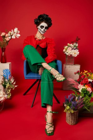 woman in dia de los muertos makeup sitting in armchair near traditional altar with flowers on red