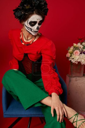Frau in Catrina Make-up sitzt im Sessel neben Blumen auf rot, dia de los muertos tradition