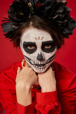 woman in spooky skull makeup and black wreath looking at camera on red, dia de los muertos concept