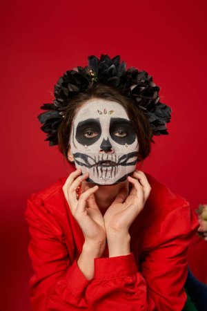 espeluznante mujer en maquillaje de cráneo y corona negra cogida de la mano cerca de la cara en rojo, día de los muertos