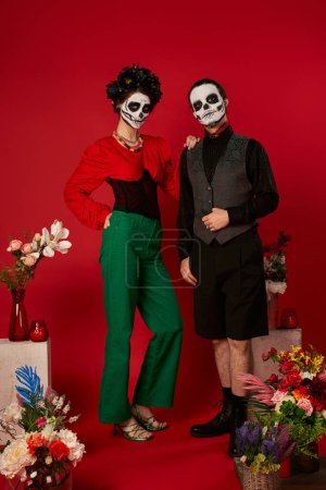 Téléchargez les photos : Couple élégant dans le maquillage du crâne de sucre près traditionnelle dia de los muertos ofrenda avec des fleurs sur rouge - en image libre de droit