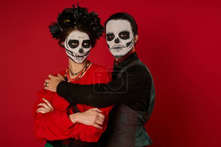 couple in dia de los muertos makeup, man embracing woman standing with folded arms on red