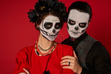 Stilvolles Paar in gruseliger Zuckerschädel-Schminke mit Blick in die Kamera auf dem roten, dia de los muertos festival