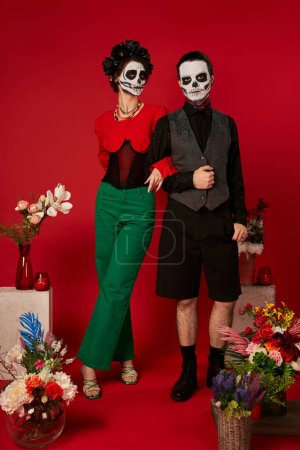 Paar in Zuckerschädel-Make-up in der Nähe des traditionellen dia de los muertos Altars mit Blumen auf rotem Hintergrund