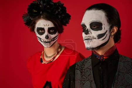 Téléchargez les photos : Dia de los muertos couple, femme dans le maquillage du crâne et couronne noire regardant caméra près de l'homme sur rouge - en image libre de droit
