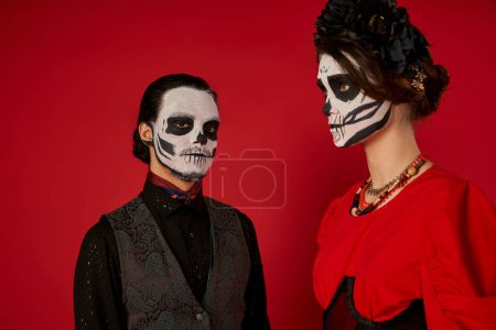 Téléchargez les photos : Homme dans le crâne maquillage regarder caméra près de la femme en couronne noire, dia de los muertos couple sur rouge - en image libre de droit