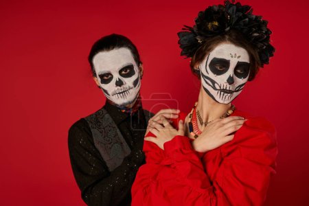 woman in skull makeup and black wreath posing with crossed arms near spooky man on red, Day of Dead