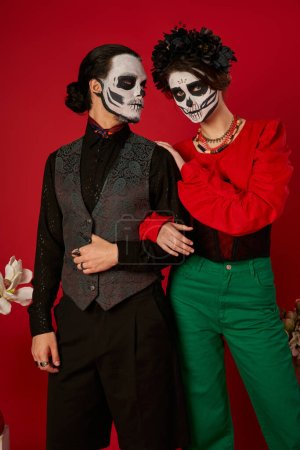 Téléchargez les photos : Dia de los muertos couple, femme dans le maquillage du crâne et couronne noire debout près de l'homme étrange sur rouge - en image libre de droit