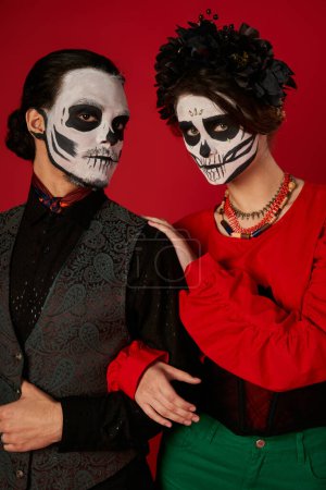 Téléchargez les photos : Couple effrayant dans le maquillage catrina calavera regardant la caméra sur rouge, dia de los muertos tradition - en image libre de droit