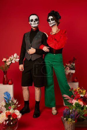 full length of couple in skull makeup near traditional Day of Dead ofrenda with flowers on red