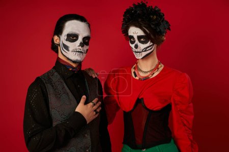 Gruseliger Mann mit Hand auf der Brust neben Frau in Zuckerschädel-Make-up, dia de los muertos Paar auf rot
