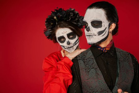 woman in catrina makeup and black wreath leaning on shoulder of eerie man on red, Day of Dead