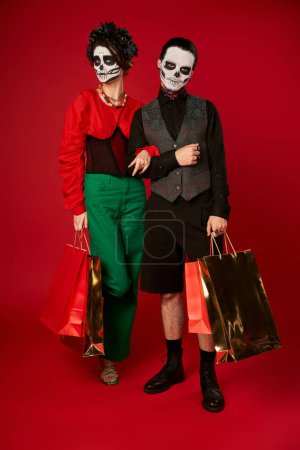 full length of trendy couple in sugar skull makeup holding shopping bags on red, dia de los muertos