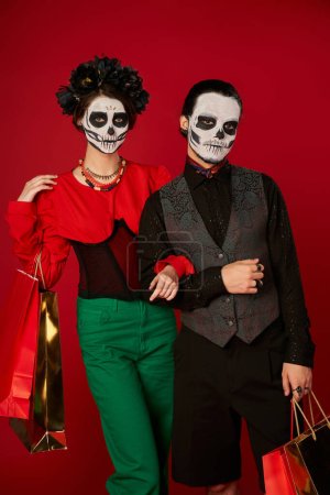 stylish couple in dia de los muertos makeup standing with shopping bags on red, seasonal sale