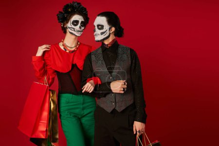 spooky couple in dia de los muertos makeup standing with shopping bags on red, festive seasonal sale