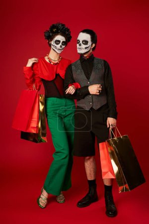 Téléchargez les photos : Longueur totale du couple dans dia de los muertos maquillage debout avec des sacs à provisions sur rouge, vente festive - en image libre de droit
